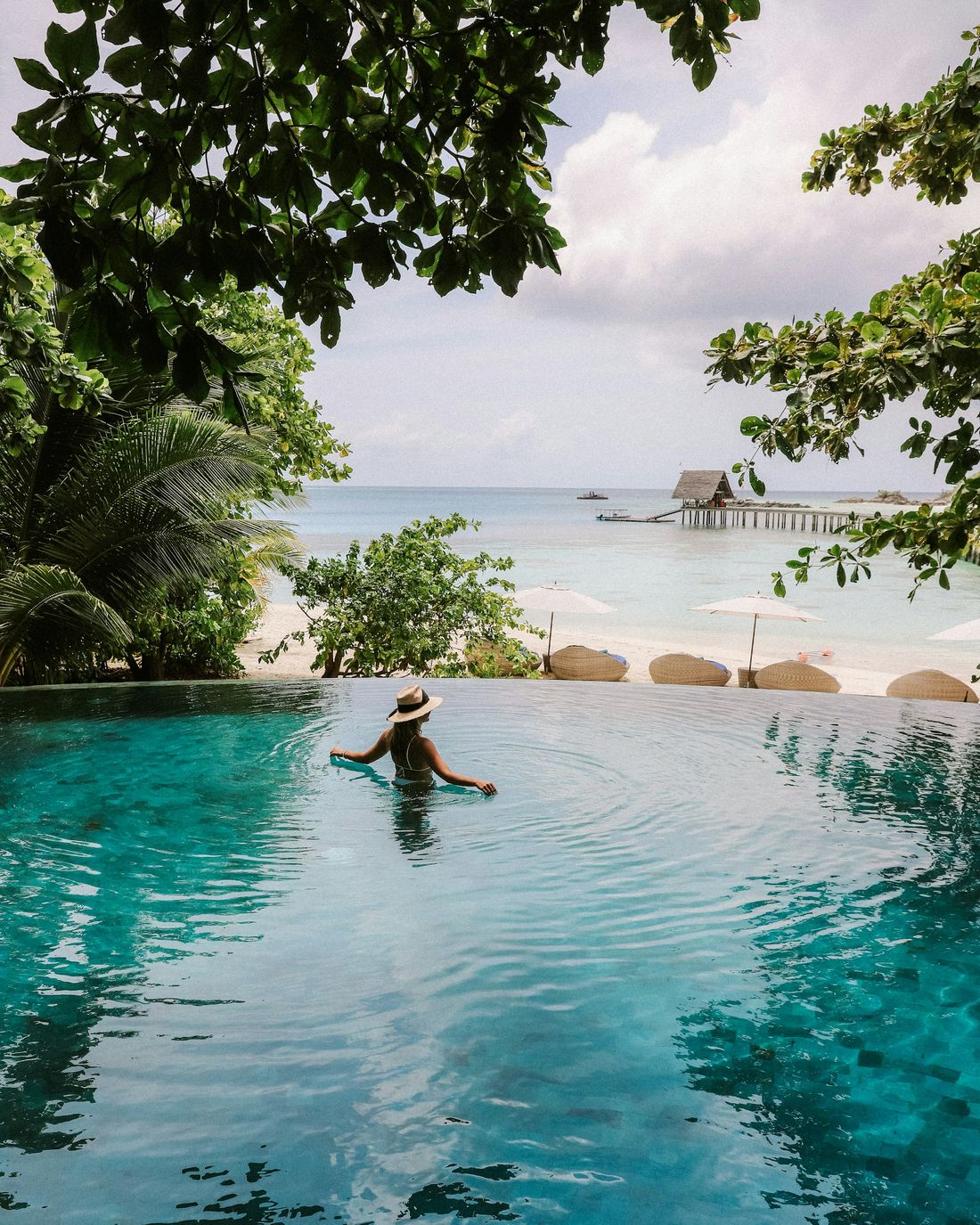 A traveler in a straw hat in an infinity pool overlooking a white-sand beach and turquoise sea